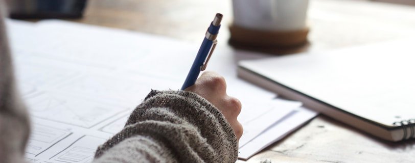 person writing on brown wooden table near white ceramic mug