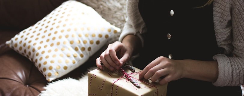 woman in grey sweater and blue denim dungarees sitting on brown and white sofa opening a yellow gift box closeup photography