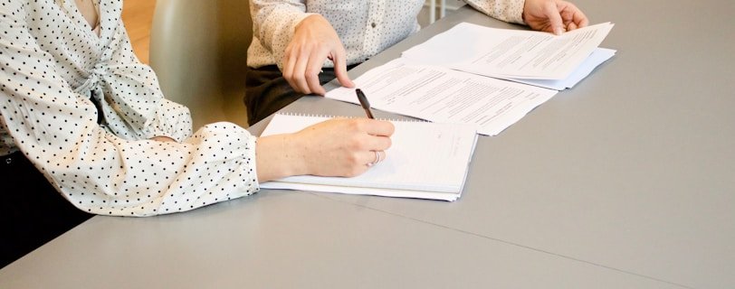 woman signing on white printer paper beside woman about to touch the documents