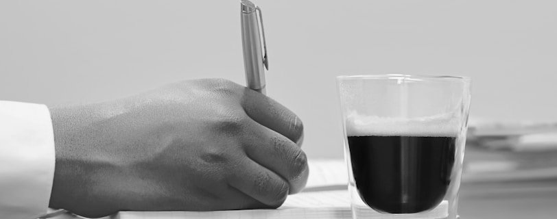 person writing on note book beside filled cup